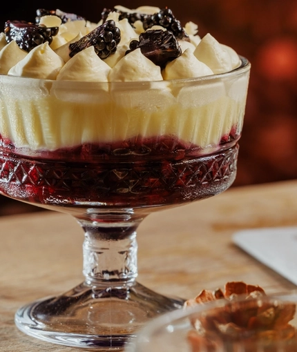 An advertising image of a Trifle dessert and a glass of white wine within the interior restaurant seating area at The Foundry Bell.