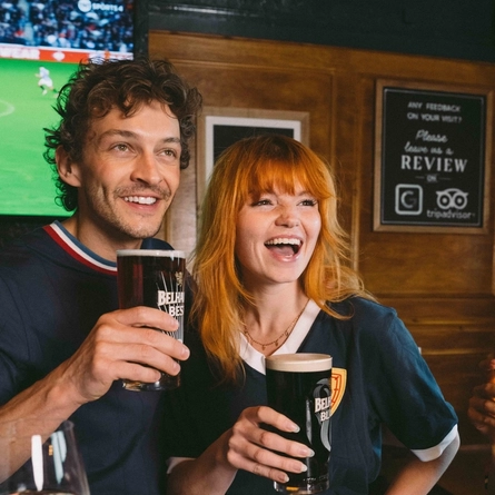 An image focusing on 2 people within a group of friends stood at a table watching a football match and enjoying a selection of drinks and snacks within the interior seating area at a Greene King venue.