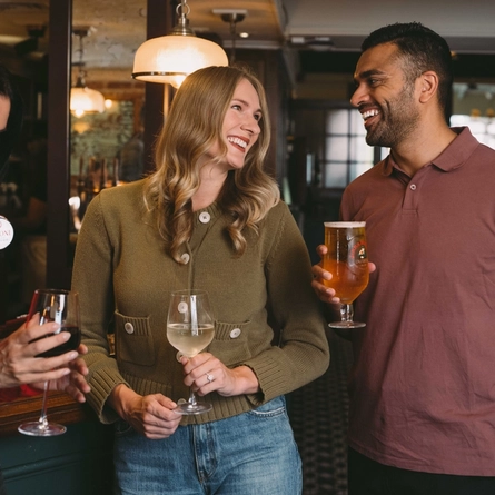 Three people standing at a pub bar holding a glass of red wine, a glass of white wine and a pint of beer.