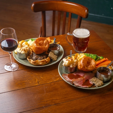 Two plates of roast dinner, one with Mushroom Tart and the other with Roast Beef, served on plates which sit on a wooden restaurant table along with a glass of red wine and a glass of beer.