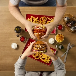 A top down view of two people sitting at a wooden table, each holding a burger. On the table are plates of fries and onion rings, along with condiments, cutlery, drinks, and small pots of dipping sauces.