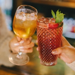 A cocktail served in a wine glass and topped with mini marshmallows, and another cocktail served in a highball glass and garnished with mint, being clinked together inside a pub bar area.