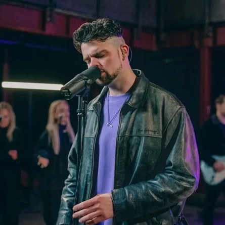A man wearing a black leather jacket, singing into a microphone on a stand.