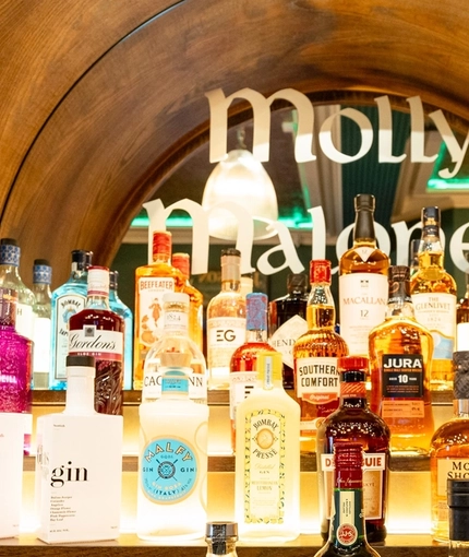 A close up view of glasses and three bottles of different whiskeys sitting on a counter at Molly Malones, with bottles of other whiskeys and gins sitting on illuminated shelves behind them.