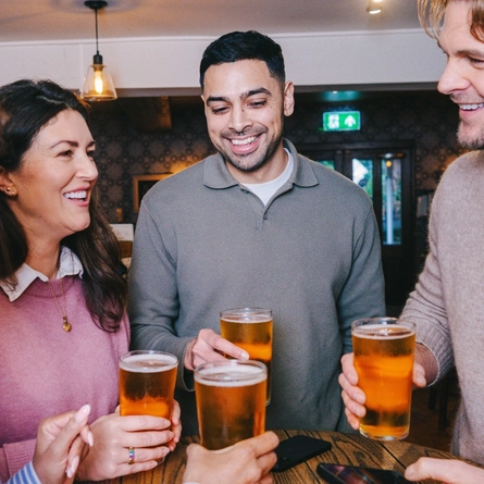 Four people gathered around a small round table inside a pub, each of them holding a glass of cider.