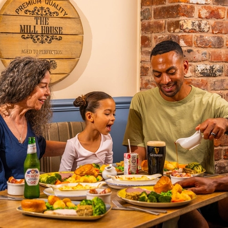 Three people seated around a wooden table in a pub, sharing Sunday roast meals. One person pours gravy from a small jug onto a plate of roast dinner with Yorkshire puddings, vegetables and potatoes. A bottle of alcohol-free beer, a pint of Guinness and a children's juice carton sit among the dishes. The background features blue panelled walls, exposed brickwork and a wooden barrel-style sign reading “The Mill House”.
