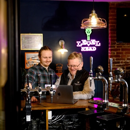 Two Men stood at the bar looking at a laptop and smiling