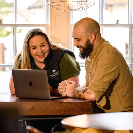 A Woman and Man sat at a table looking at a laptop and smiling