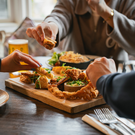A wooden serving board laden with a variety of food, sitting on a restaurant table next to cutlery, a glass of beer, and other plates of food. Three people at the table are picking up pieces of food from the serving board.