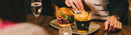 A person sitting at a restaurant table with a burger and a small bowl of chips served on a plate in front of them. A glass of champagne and a Christmas cracker also sit on the table, and a Christmas tree is visible in the background.