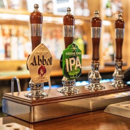 A close up view of the beer taps for Abbot Ale and Greene King IPA, on a bar inside a pub.