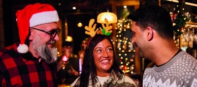 Three people are standing at a tall table with drinks,  in a festive, warmly lit bar. Two of them are wearing Christmas themed headwear.  The background features Christmas decorations, including garlands and lights, as well as hanging glasses above the bar area.