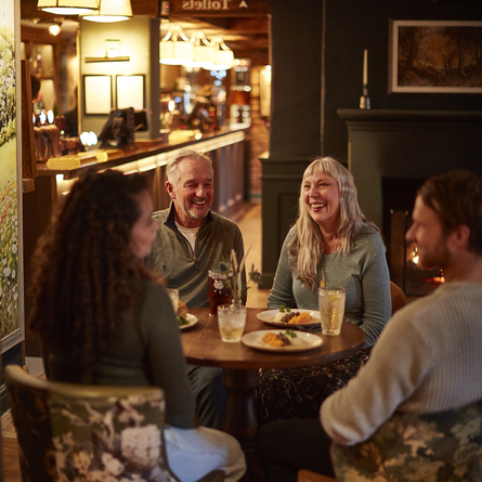 4 people sat around a table with drinks. In front of the fire and the bar.
