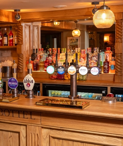 A close up view of the wood panelled bar inside the Ye Olde Leathern Bottel in Barkham, with spherical glass lights hanging above the counter, and bottles of wine and alcoholic spirits on wooden shelves behind the bar.