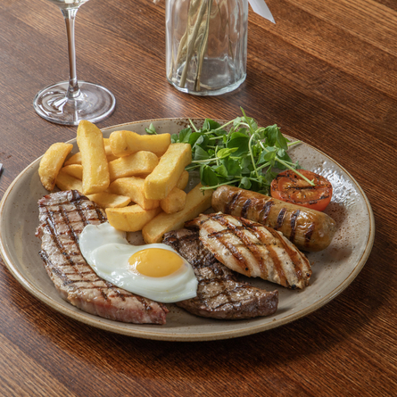 A mixed grill meal at Chef & Brewer venues. The meal features steak, fried egg, chicken, sausage, tomato, chips and salad served on a plate which sits on a wooden table.