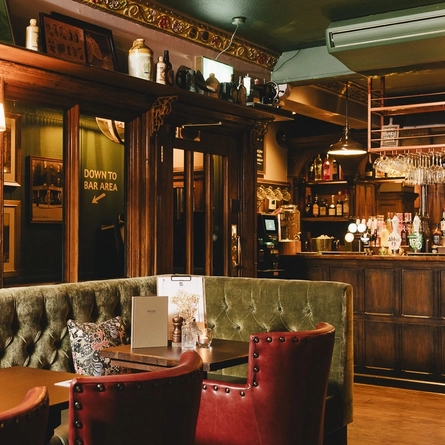 The wood panelled bar and interior restaurant seating area at the Masons Arms, with upholstered booth seats, ornaments on a wooden shelf, and flowers carved into the coving.