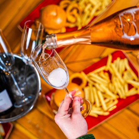 Prosecco being poured into a champagne flute which is being held above a table laden with plates of burgers and chips.