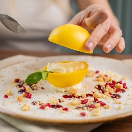 A close up view of a Lemon Cheesecake dessert in a yellow, lemon shaped shell, served on a plate. The plate sits on a cloth napkin on a wooden table, and a person at the table is holding a spoon in one hand while removing the top half of the lemon shell with the other hand.