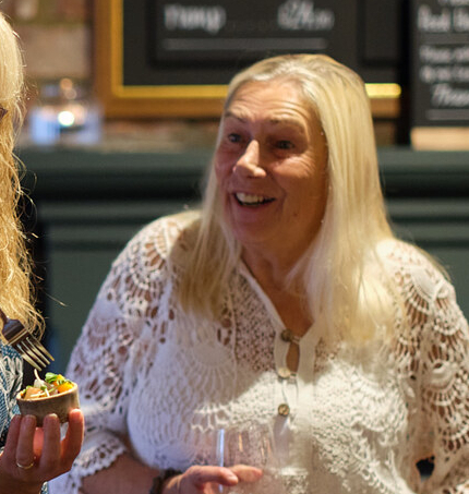 A group of guests inside the Smiths Arms pub, one person is holding a drink in a glass and another person is using a fork to sample food from a small pot they're holding.