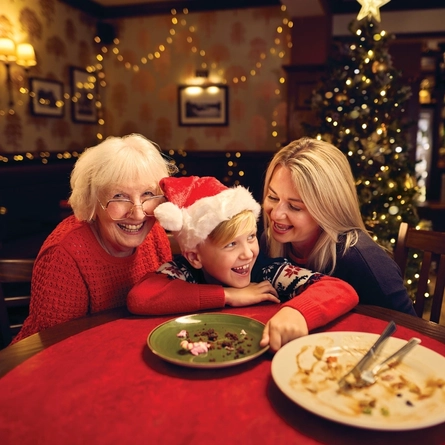 A child and two women sitting at a table inside a pub, empty plates and cutlery sitting on the table in front of them. A Christmas tree and fairy lights are visible in the background.