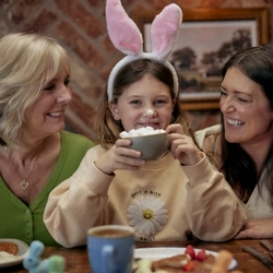 An image of a child wearing bunny ears  smiling at the camera and enjoying a hot chocolate.