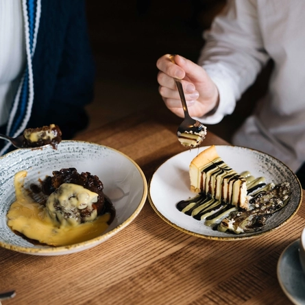 A close up view of a bowl of pudding and custard, and a plate of cheesecake, sitting on a wooden restaurant table along with two cups of coffee on saucers. Two people sitting at the table are holding spoons with scoops of the desserts on.