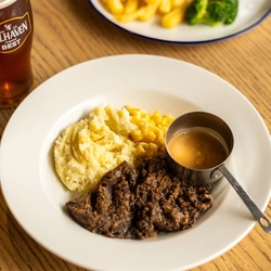 Haggis, Neeps and Tatties served on a plate with a small pot of sauce. The plate sits on a wooden table next to a glass of Belhaven Best ale and another plate of food.