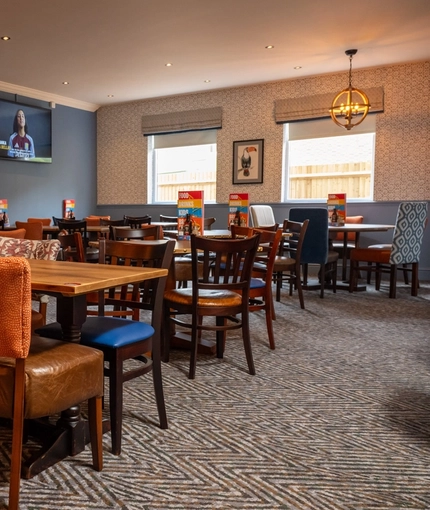 The interior restaurant seating area at The Heron in Havant; with wooden tables and chairs, a blue velvet booth seat against one wall, two windows at the far end of the room, and a TV on the wall.