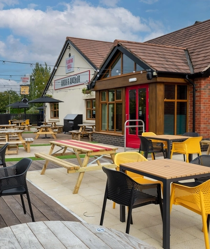 The exterior facade and beer garden seating area at the Arch & Anchor, with wooden picnic tables and string lights. 