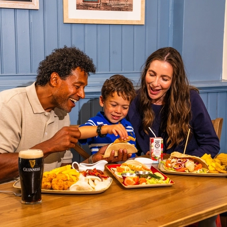 Two adults and a child are seated at a wooden table in a pub, enjoying a midweek meal. The table is filled with dishes including tacos, chips, corn on the cob and breaded bites, alongside a pint of Guinness, a glass of cola with lemon and a children's juice carton. Blue panelled walls and wooden seating create a relaxed pub setting in the background.