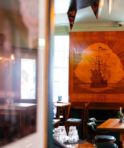 An interior restaurant seating area at the Royal George; with a TV on the wall, a fireplace, and a large wooden inlay picture of a sailing ship on the wall above it.