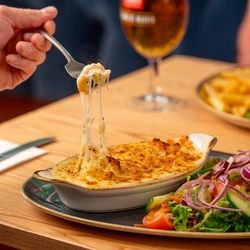 A roasting dish filled with Mac & Cheese, served on a plate with salad. The plate sits on a wooden restaurant table along with a glass of beer and another plate of food, and a hand is using a fork to pick up some of the melty mac & cheese.