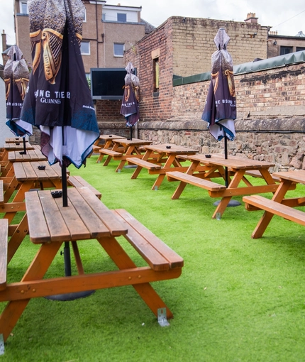 The exterior beer garden seating area of The Merlin in Edinburgh, with wooden picnic benches and shade umbrellas.