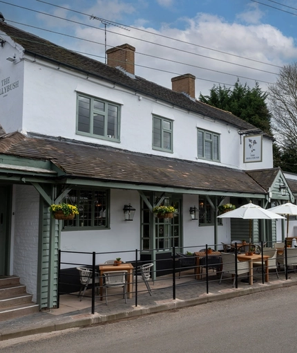 The exterior facade, signage, and seating area of The Hollybush in Lichfield, with shade umbrellas on some of the wooden tables, and flower baskets hanging from the veranda.