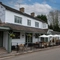 The exterior facade, signage, and seating area of The Hollybush in Lichfield, with shade umbrellas on some of the wooden tables, and flower baskets hanging from the veranda.