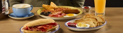 Breakfast dishes at Hungry Horse venues, including a plate of sausages, fried egg, bacon, baked beans, mushroom, and hash browns, a plate of slices of toast with butter, and another plate holding a breakfast sandwich. The plates sit on a wooden table along with a cup of coffee, a glass of orange juice, condiments, and a small pot of dipping sauce.