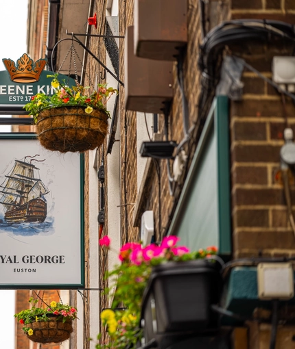 A close up view of the pub sign hanging in between two flower baskets on the exterior wall of the Royal George. The sign features an illustration of a large ship sailing on the sea above the words "Royal George, Euston", on a white background.