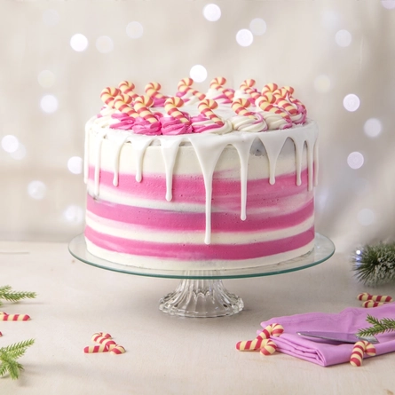 The Candy Cane Lane cake served on a glass cake stand which sits on a table next to a napkin and cake slicer. The table is scattered with pine tree sprigs and mini candy cane decorations.