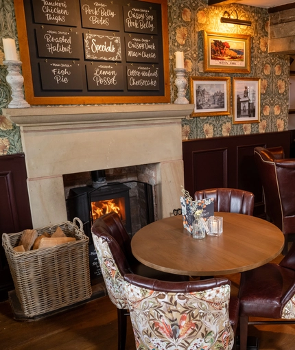 An interior restaurant seating area of The Hollybush, with upholstered chairs, wooden beams, and a fireplace.