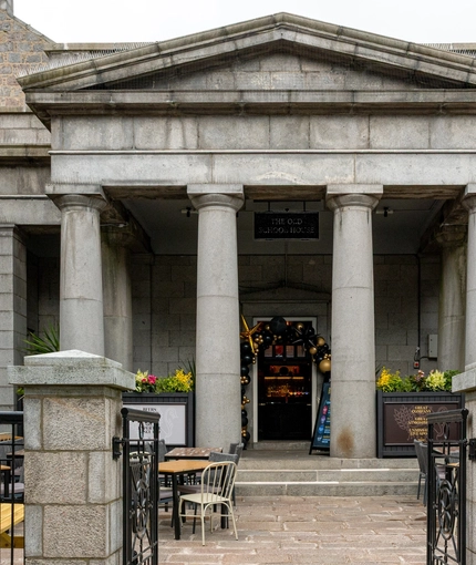 The exterior facade, signage, and beer garden seating area of the Old School House in Aberdeen, with large stone columns, planter boxes, and wooden picnic tables.
