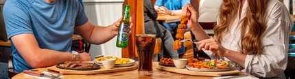 Two people sitting at a wooden restaurant table. One person has a plate of steak and chips on the table in front of them and is holding a bottle of Heineken 0.0% alcohol beer; the other person is holding a skewer of chicken and is using a fork to remove a piece. A glass of Coca-Cola sits on the table next to them.