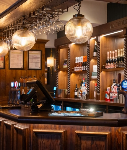The wood panelled bar at The Malthouse Farm in Whittle-Le-Woods, with wine glasses and spherical lights hanging above the counter, and bottles of wine on wooden shelves behind the bar.