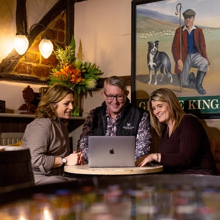 Two women sit on either side of a man as they look at a laptop on the table smiling 