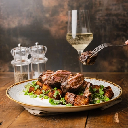 A close up view of a plate of Lamb Rump served with potatoes and peas. The plate sits on cloth napkin on a wooden table along with salt and pepper mills, and a glass of sparkling wine; and a hand is holding a fork with a piece of lamb on it above the plate.