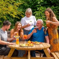 A mixed group of people seated and stood around a wooden table, in a beer garden, enjoying a variety of sharers and drinks.