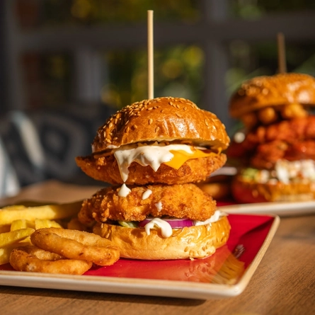A close up view of a Smokey Rancher Burger served on a red plate with fries and onion rings. The plate sits on a sunlit wooden table along with other plates of food.