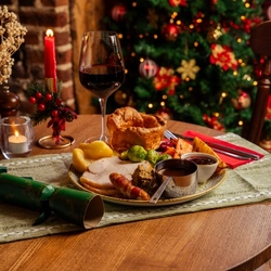 A Traditional Turkey Dinner served on a plate, featuring Yorkshire pudding, roast potatoes, vegetables, sliced turkey, stuffing, pigs in blankets, and pots of gravy and cranberry sauce. The plate sits on a wooden restaurant table along with cutlery, a serviette, salt and pepper mills, a Christmas cracker, table decorations, and a glass of red wine; and a Christmas tree is visible in the background.