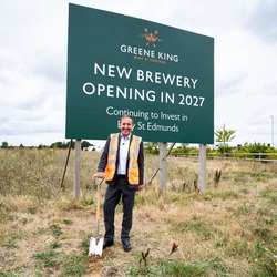 Nick Mackenzie in a hi vis vest, stood with a shovel in front of a sign announcing the start of the new brewery being built.