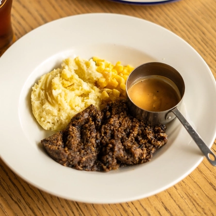 Haggis, Neeps and Tatties served on a plate with a small pot of sauce. The plate sits on a wooden table next to a glass of Belhaven Best ale and another plate of food.