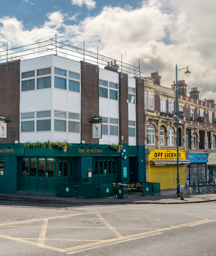 The dark green exterior facade, golden yellow signage, and exterior seating area at The Junction in Upminster, with a view of the neighbouring buildings.